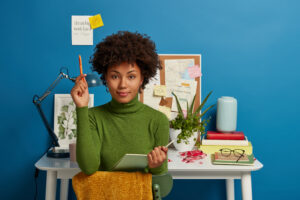 Serious African American student makes plan of preparation for exam, writes in notepad, makes notes, poses against cozy interior at workplace, white desk with lamp, glasses, papers, blue background